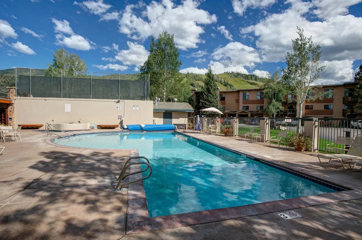 A crystal-clear outdoor swimming pool surrounded by tall trees and lounge chairs, with a dramatic mountain backdrop visible in the distance. The tranquil setting reflects the resort’s relaxation amenities after a day of hiking or biking.