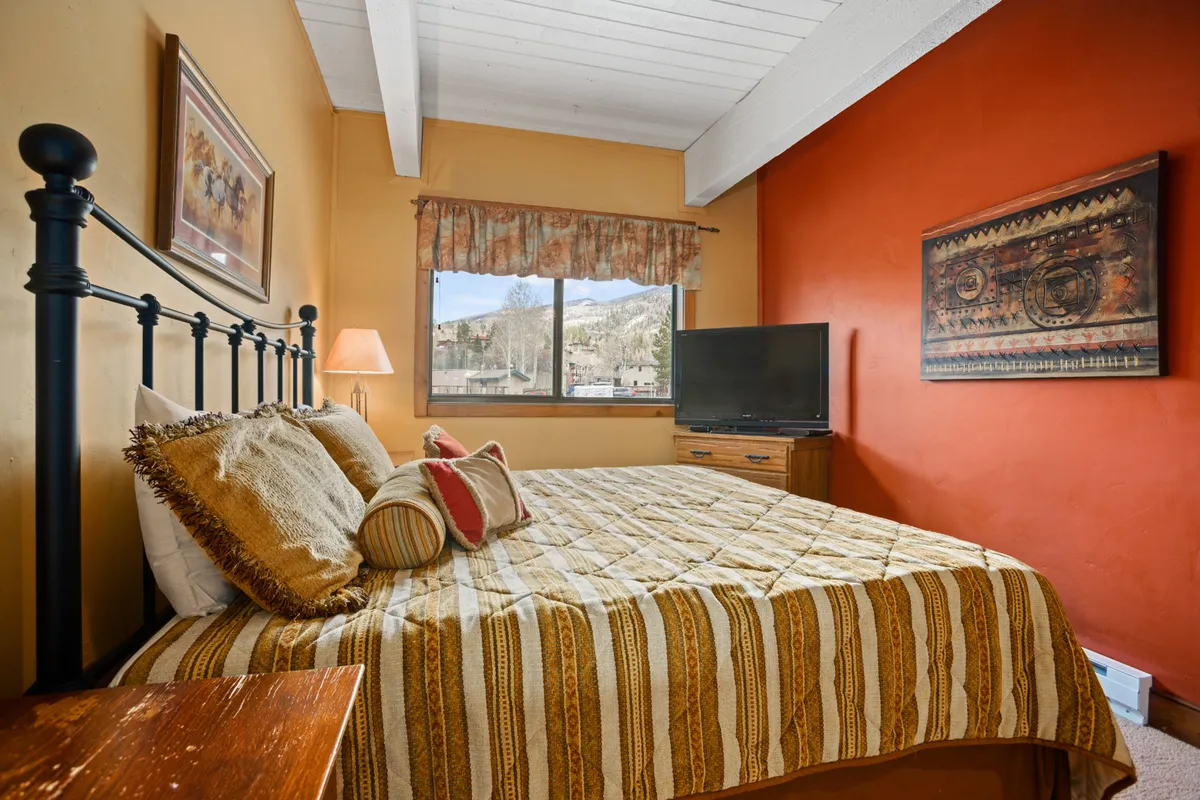 This interior bedroom scene centers on a bed with patterned bedding, positioned next to a window that frames a dramatic mountain view. The cozy, relaxed setting makes it easy to imagine waking up to the alpine landscape, further connecting interior comfort with the outdoor scenery.