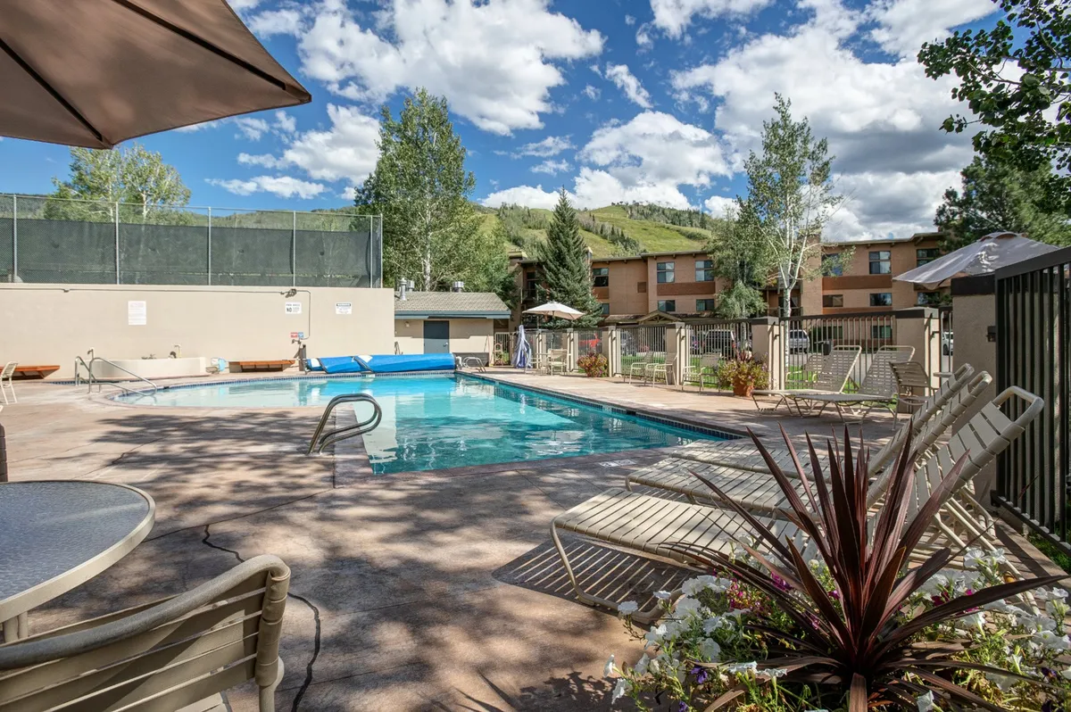 An inviting pool area framed by trees, dotted with lounge chairs and shaded by umbrellas, with the colorful forest providing a lush, summer atmosphere. The scene captures the leisure side of the property, perfect for post-activity relaxation.