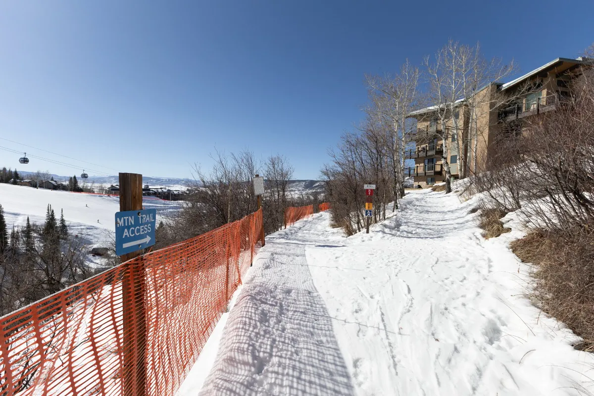 A snow-covered pathway leads directly to a mountain trail access sign, with surrounding trees and the building structure visible in the background. The scene is set for winter activities, with the environment inviting families to begin their ski day just steps from their accommodation.
