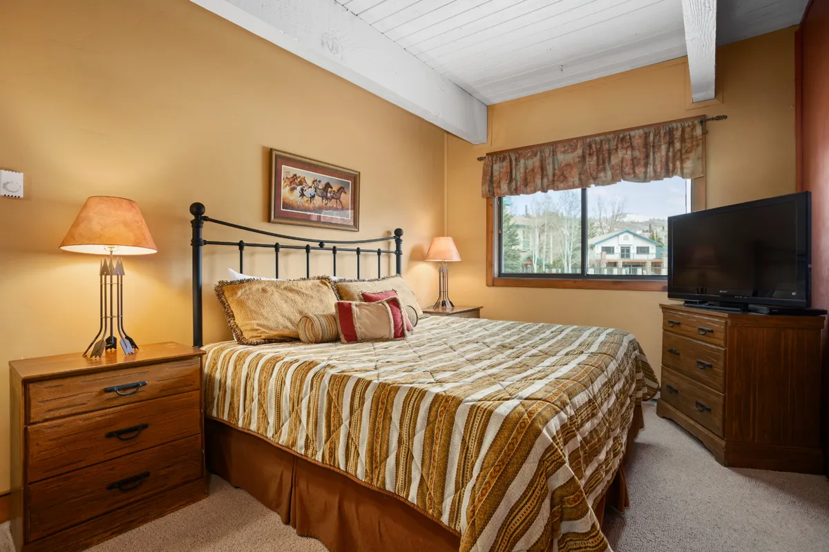 A spacious bedroom featuring a king-sized bed with patterned bedding, a nightstand with a lamp, and a large window offering mountain views. The room also includes a television for evening relaxation. The soft lighting and inviting decor create a tranquil, restful atmosphere.