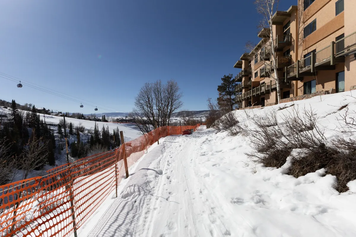 This image captures gondola lifts gliding through the sky above a snow-covered pathway leading toward a welcoming building facade, with trees lining the route. The scene embodies the vibrancy of Steamboat’s après-ski atmosphere and the journey from the slopes to the heart of social activity.