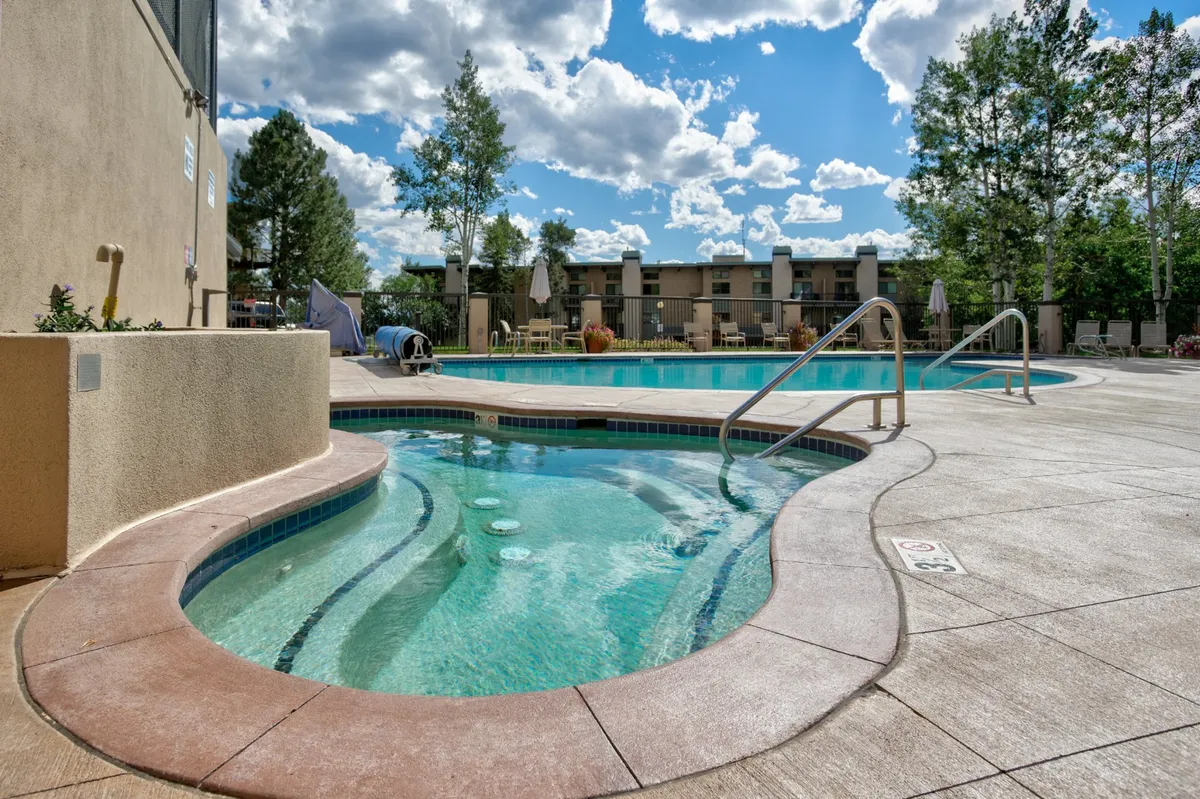 This image features a resort-style outdoor area with a hot tub and swimming pool surrounded by lounge chairs and trees, under an open sky. The tranquil setting showcases both the pool and hot tub, inviting guests to relax, sunbathe, or socialize poolside.