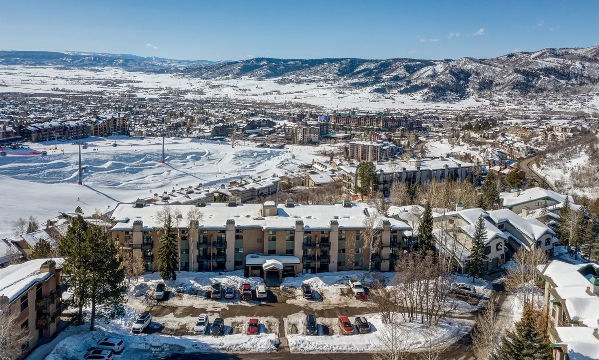 An elevated view of snow-covered rooftops set against surrounding mountains, with adjacent buildings and a visible parking area below. The scene captures the development's proximity to both natural beauty and neighboring condos—ideal for group cohesion and easy movement between units.