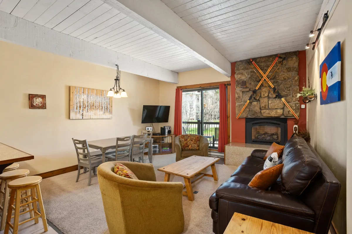A welcoming open-concept living and dining area featuring a gas fireplace as the focal point. The space includes a large dining table for six, comfortable seating, and expansive windows that offer ample natural light and views of the surrounding landscape. The wood-paneled ceiling and exposed beams add a touch of mountain lodge charm, while tasteful artwork completes the cozy vibe.