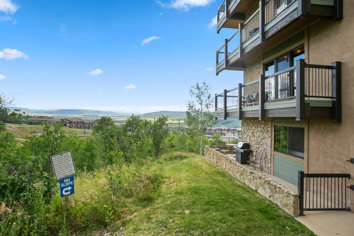 This image features a balcony with railings overlooking a lush green lawn, trees, and a visible ski slope sign in the distance. The balcony is partially covered, offering space for relaxing or dining while enjoying the outdoors. The scene suggests opportunities for both quiet morning coffees and evening sunset dinners, with the mountains offering a scenic backdrop.