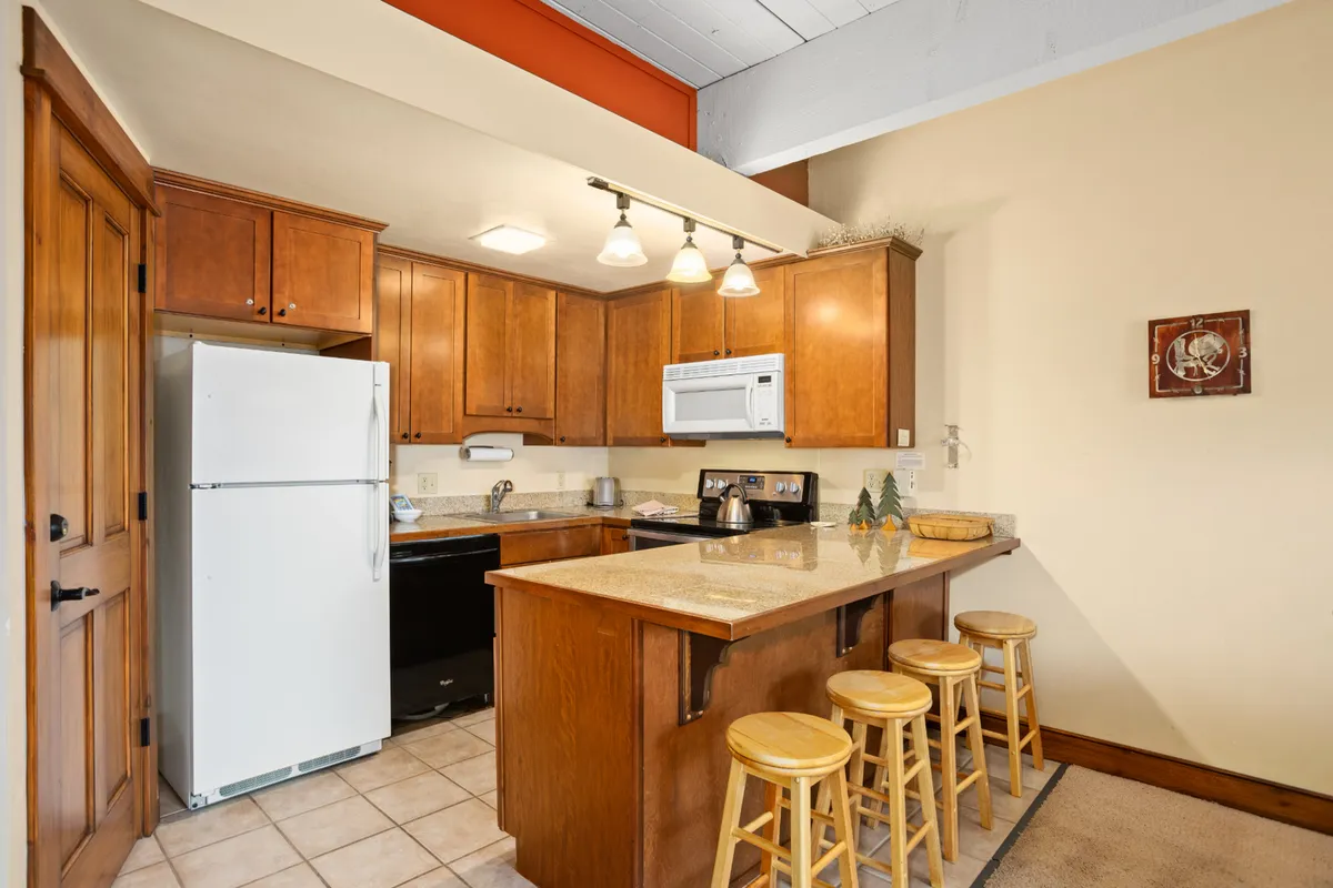 A modern kitchen with granite countertops, wooden cabinetry, a kitchen island equipped with bar stools, and stainless steel appliances. The layout is open to the main living space, allowing for easy interaction during meal preparation and casual dining.