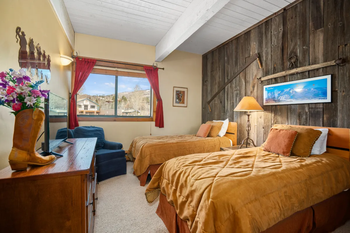 A cozy second bedroom with two twin beds, a rustic barnwood accent wall, a window offering outside views, a nightstand with a lamp, and a decorative flower arrangement. The layout is perfect for children, friends, or solo travelers.